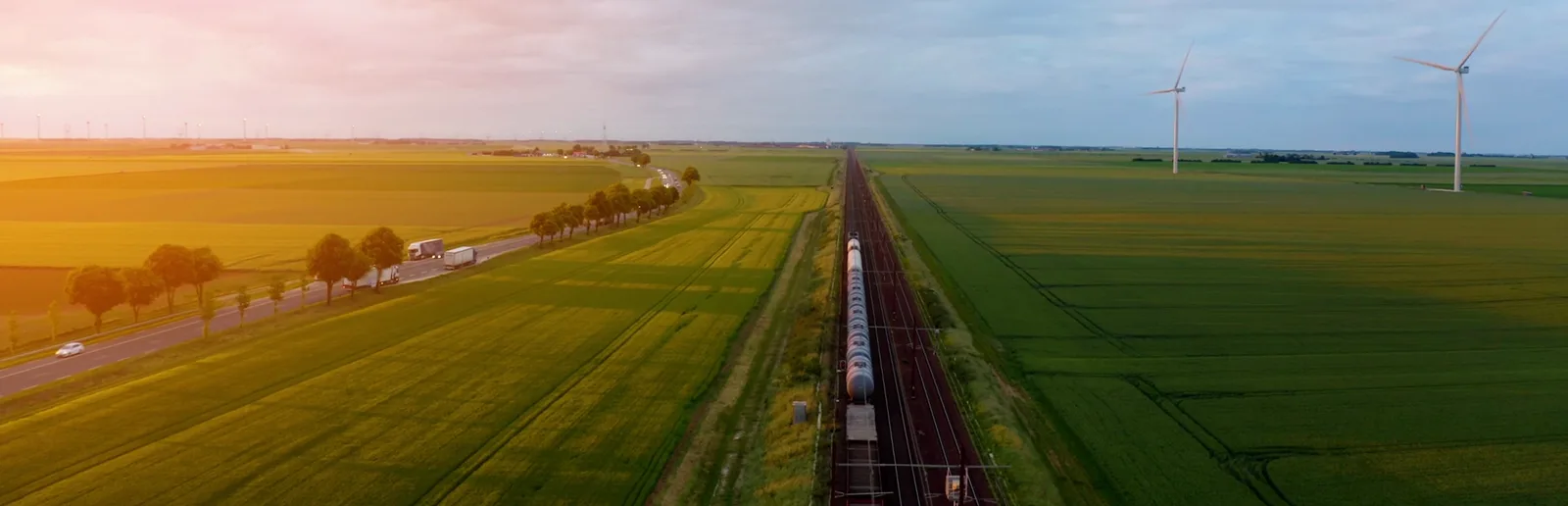 Aerial view of a freight train crossing farmland at sunset, with wind turbines on the horizon